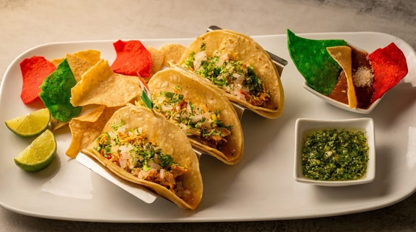 Three tacos with cilantro and onions, colorful chips, lime wedges, salsa, and green sauce.