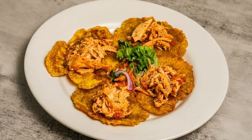 Crispy tostones topped with shredded chicken, lettuce, carrots, and red onion on a white plate.